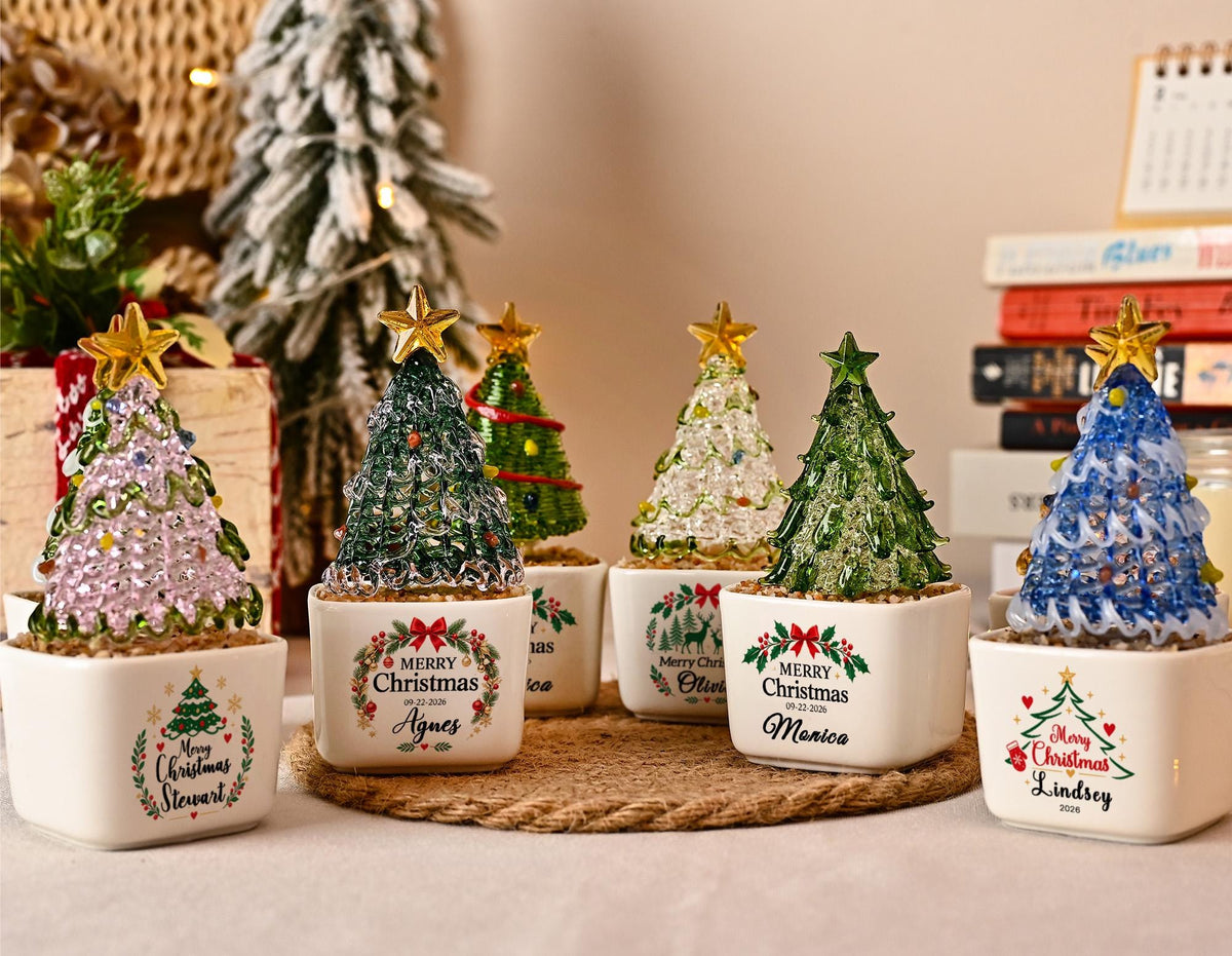 May include: A collection of six small glass Christmas tree ornaments, each set in a square white ceramic planter. The trees are various colors including pink, green, and blue, topped with gold stars. Each planter features a Merry Christmas wreath design with a personalized name printed on the front.
