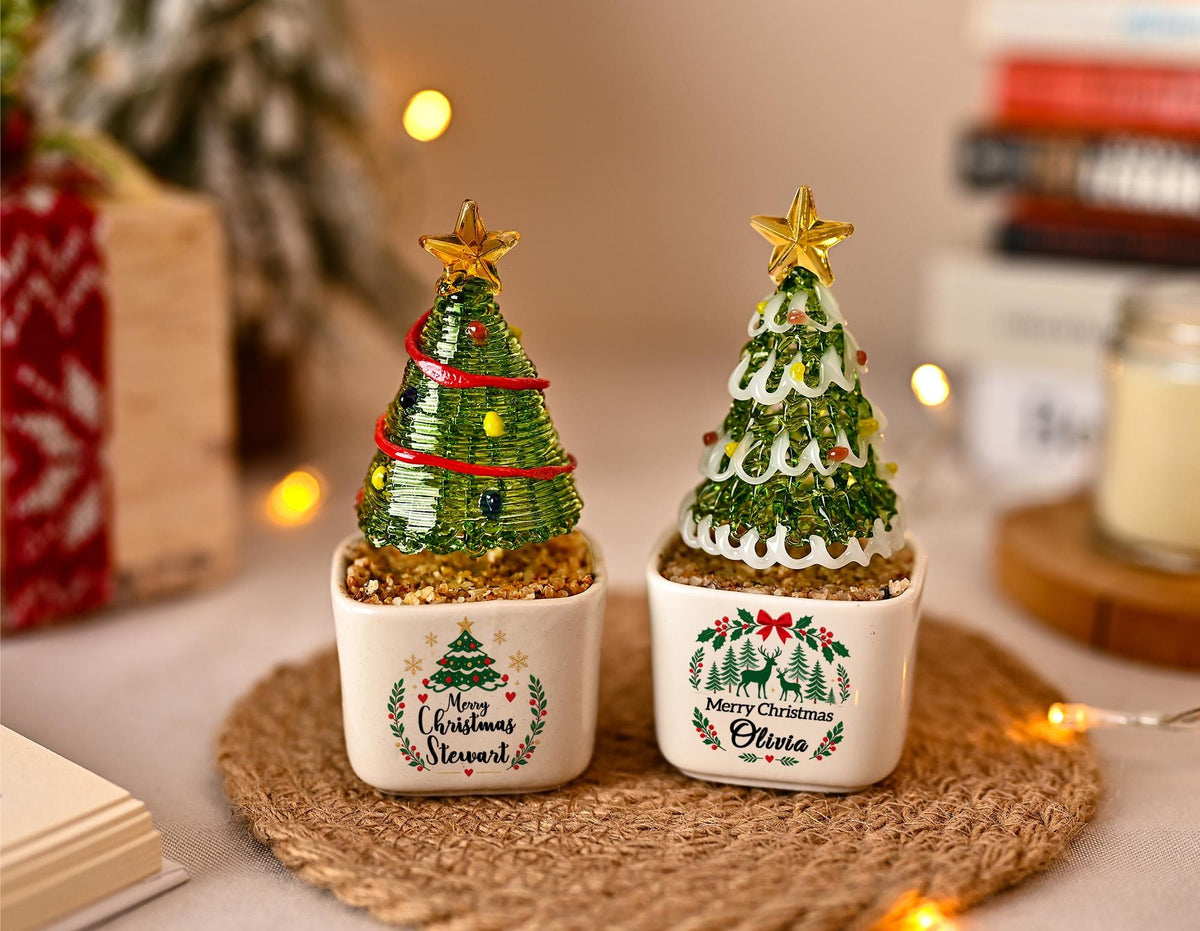 May include: Two small decorative Christmas tree figurines in square white ceramic pots. The left tree has a red garland and the text Merry Christmas Stewart. The right tree has white garland and the text Merry Christmas Olivia. Both have gold stars.