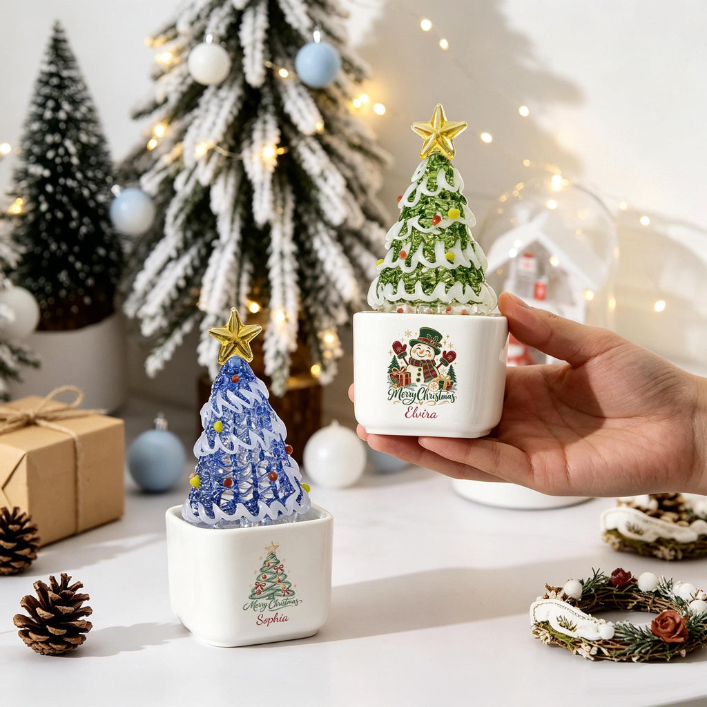 May include: Two square white ceramic containers, each holding a glass Christmas tree ornament topped with a gold star. One container features a snowman graphic with the text Merry Christmas Elvira, the other a tree with Merry Christmas Sophia.