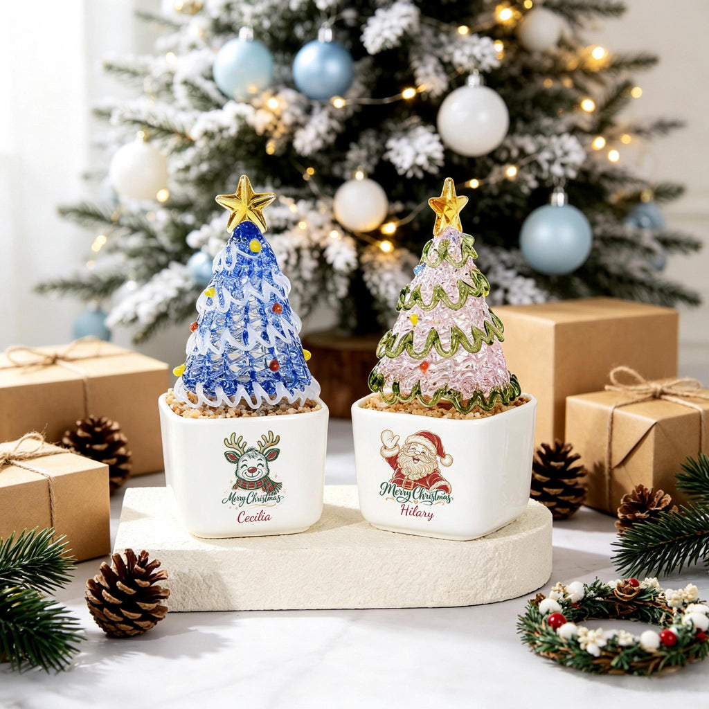May include: Two square white ceramic planters containing decorative Christmas trees. The left tree is blue with a gold star and the text "Merry Christmas Cecilia." The right tree is pink with a gold star and the text "Merry Christmas Hilary" featuring a Santa graphic.