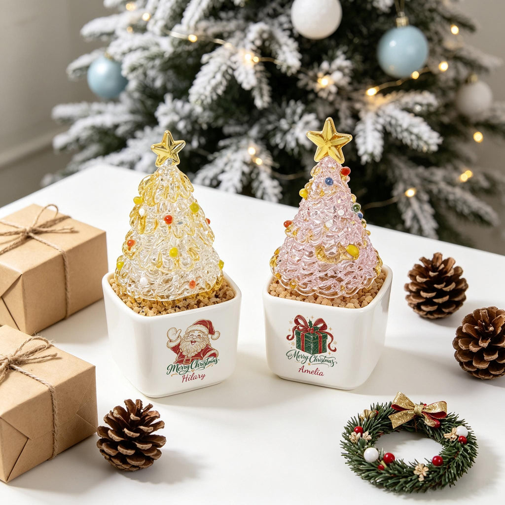 May include: Two glass Christmas tree figurines in white square ceramic pots. One tree is yellow with a gold star, the other is pink with a gold star. The pots feature Santa and gift graphics with the names Hilary and Amelia printed below.