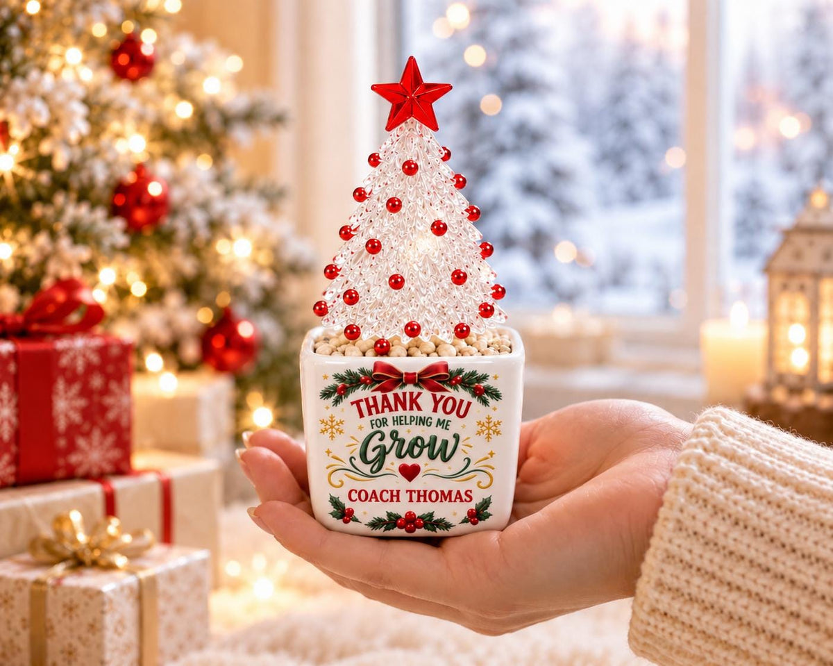 May include: A small white square ceramic planter featuring the text Thank You For Helping Me Grow Coach Thomas with a red bow and holly graphic. A clear, textured Christmas tree with red bead ornaments and a red star topper sits inside.