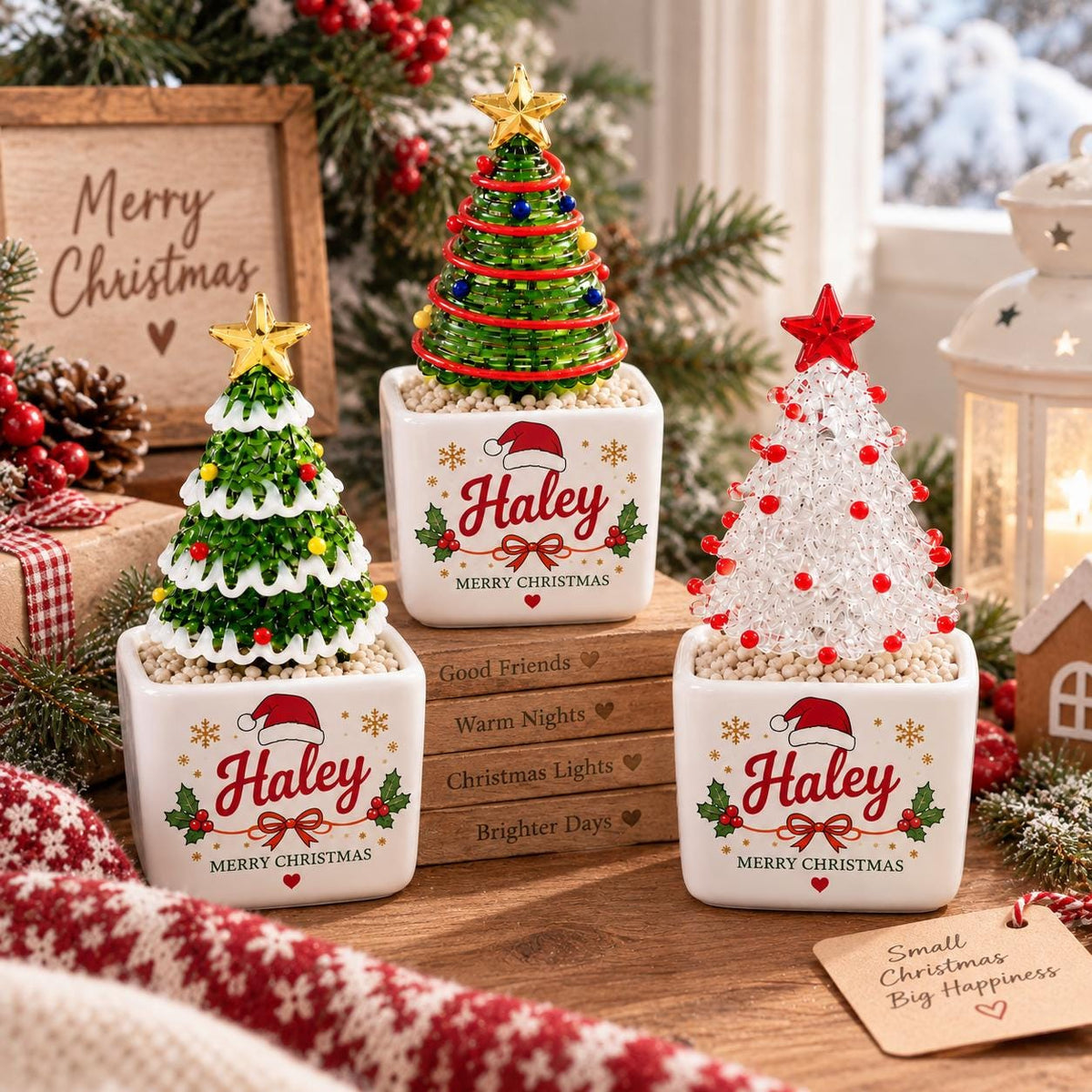 May include: Three square white ceramic planters, each containing a small decorative Christmas tree. Two trees are green with colorful ornaments and gold stars, while one is clear with red beads and a red star. Each planter features the name Haley.