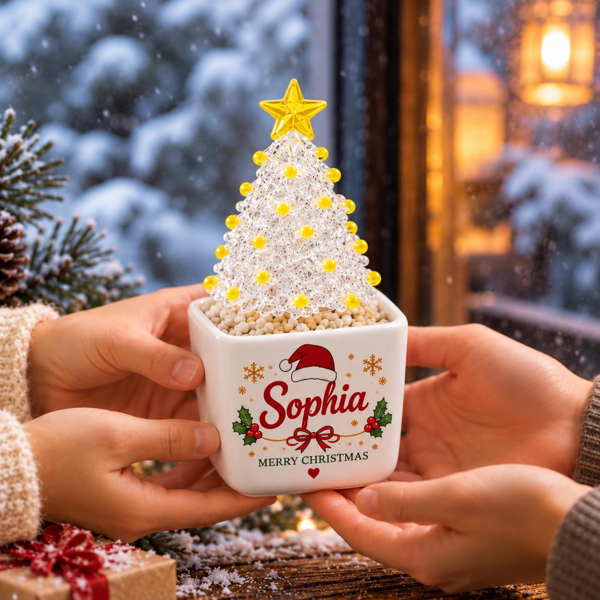 May include: A square white ceramic planter containing a clear, textured Christmas tree ornament topped with a yellow star and yellow beads. The planter features the name Sophia in red script, a Santa hat, holly, and the text Merry Christmas.