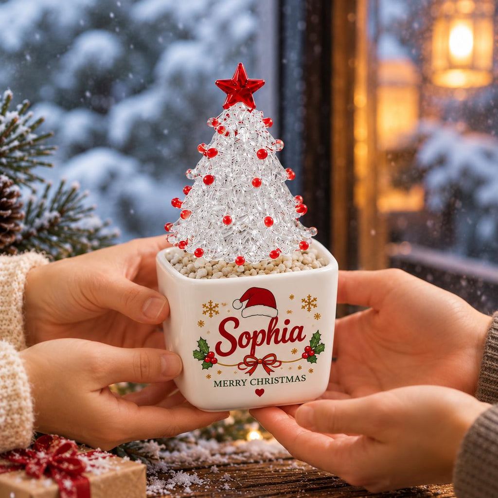 May include: A clear glass Christmas tree ornament with red bead accents and a red star topper, set in a white square ceramic planter. The planter features the name Sophia in red script, a Santa hat, holly leaves, and the text Merry Christmas.