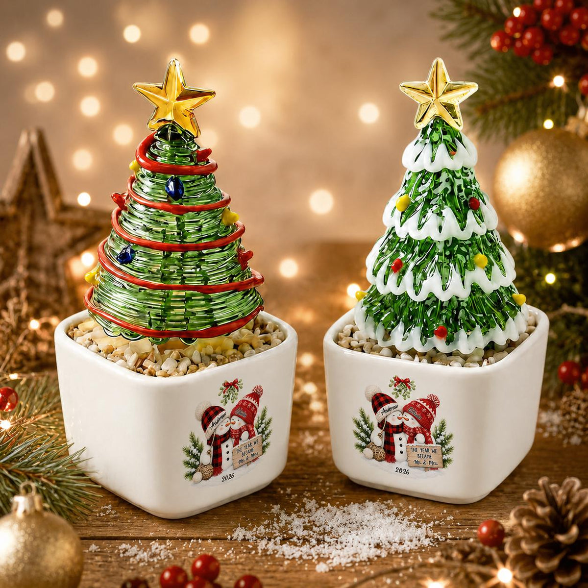 May include: Two decorative glass Christmas tree figurines in white square ceramic planters. The left tree has green glass coils with red garland and a gold star. The right tree has green glass branches with white snow and a gold star. Both planters feature a graphic of two snowmen and the text 2026.