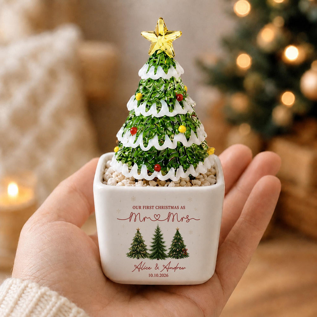 May include: A miniature Christmas tree ornament with green foliage, white snow-like trim, and small colorful baubles, topped with a yellow star. The tree sits in a white square ceramic planter printed with "OUR FIRST CHRISTMAS AS Mr & Mrs", "Alice & Andrew", and "10.10.2026" above three small tree illustrations.