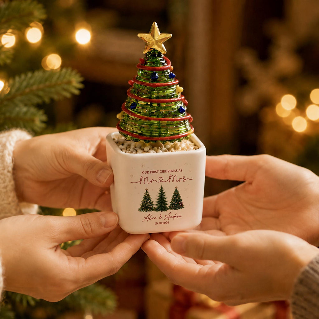 May include: A decorative glass Christmas tree ornament with a gold star topper, sitting in a white square ceramic planter. The planter features the text "OUR FIRST CHRISTMAS AS Mr & Mrs", "Alice & Andrew", and "10.10.2026" with three green pine tree illustrations.