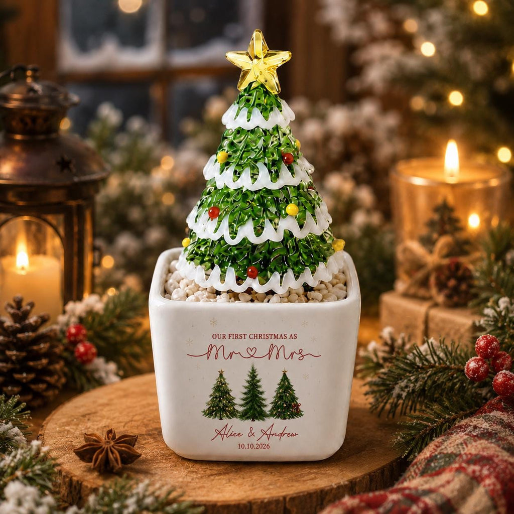 May include: A decorative tabletop Christmas tree with green branches, white snow-like trim, and a yellow star topper, set in a white square planter. The planter features the text "OUR FIRST CHRISTMAS AS Mr & Mrs", "Alice & Andrew", and "10.10.2026" with three small green tree illustrations.