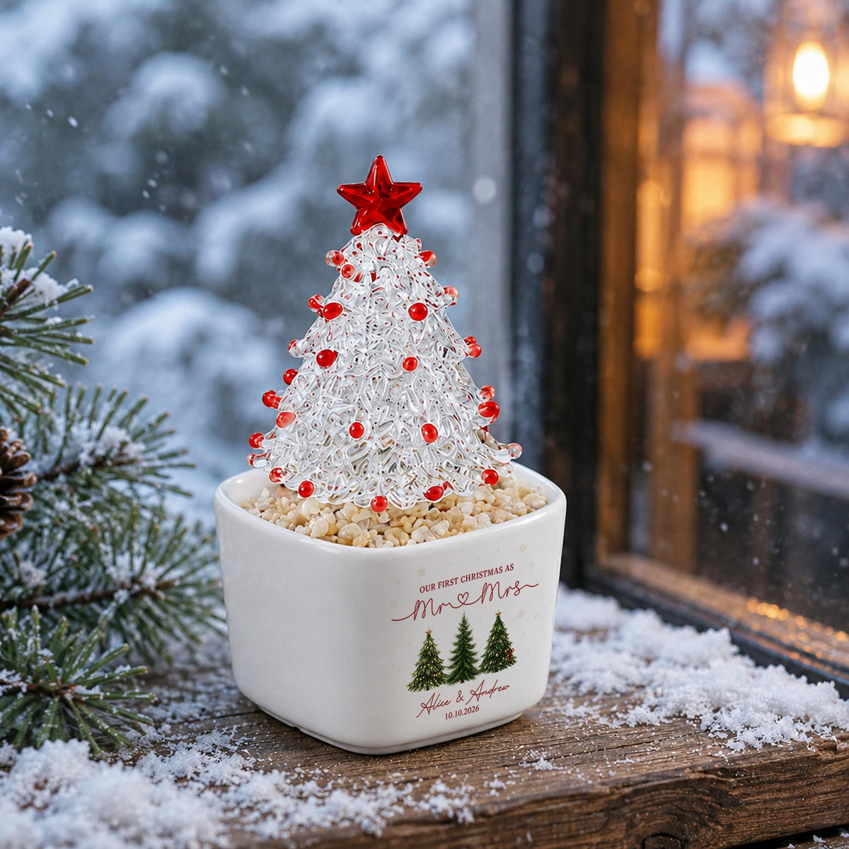 May include: A clear glass Christmas tree ornament with red glass beads and a red star topper, set in a white square ceramic planter filled with small tan pebbles. The planter features the text "OUR FIRST CHRISTMAS AS Mr & Mrs" and "Alice & Andrew 10.10.2026" with three green pine tree illustrations.