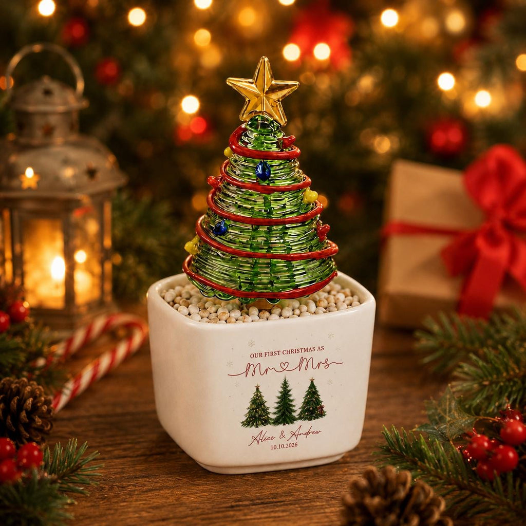 May include: A decorative glass Christmas tree with a gold star topper, red garland, and colorful ornaments, set in a white square ceramic planter. The planter features the text "OUR FIRST CHRISTMAS AS Mr & Mrs", "Alice & Andrew", and "10.10.2026" with three small green tree illustrations.