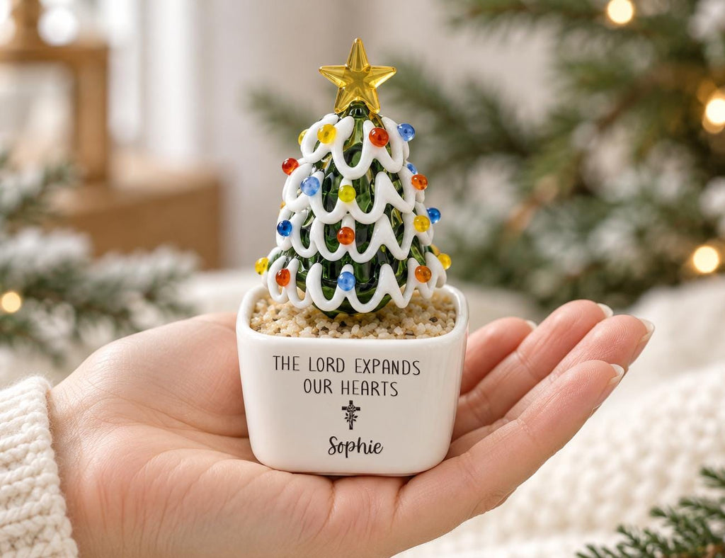 May include: A small glass Christmas tree ornament with white swirled garland and colorful glass beads, topped with a gold star. It sits in a white square ceramic planter filled with tan pebbles, inscribed with "THE LORD EXPANDS OUR HEARTS" and "Sophie".