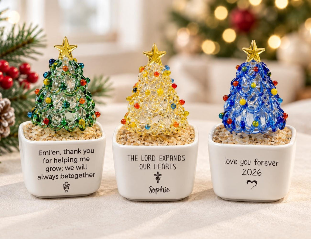 May include: Three glass Christmas tree ornaments in white square planters filled with sand. The left tree is green, the center is yellow, and the right is blue, each topped with a gold star. The planters feature printed text and symbols.