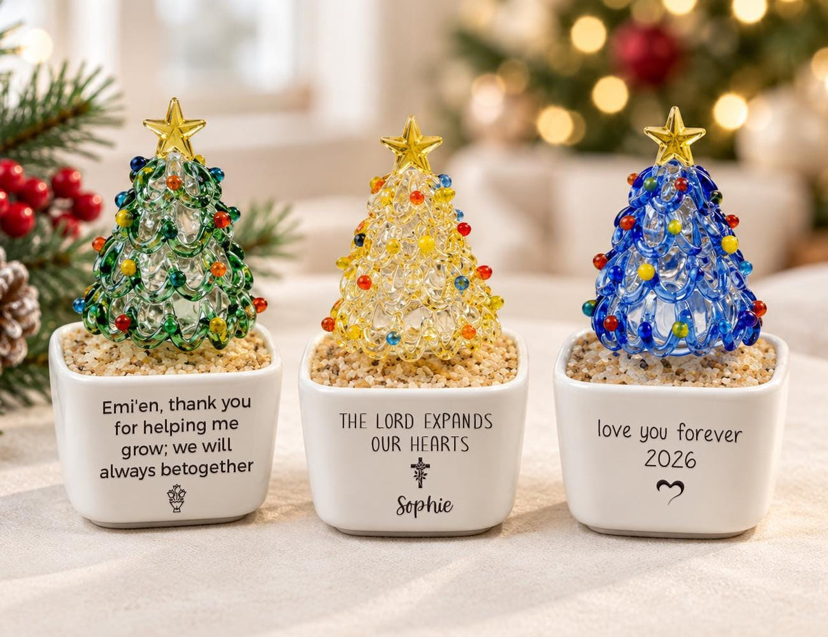 May include: Three glass Christmas tree ornaments in white square planters filled with sand. The left tree is green, the center is yellow, and the right is blue, each topped with a gold star. The planters feature printed text and symbols.