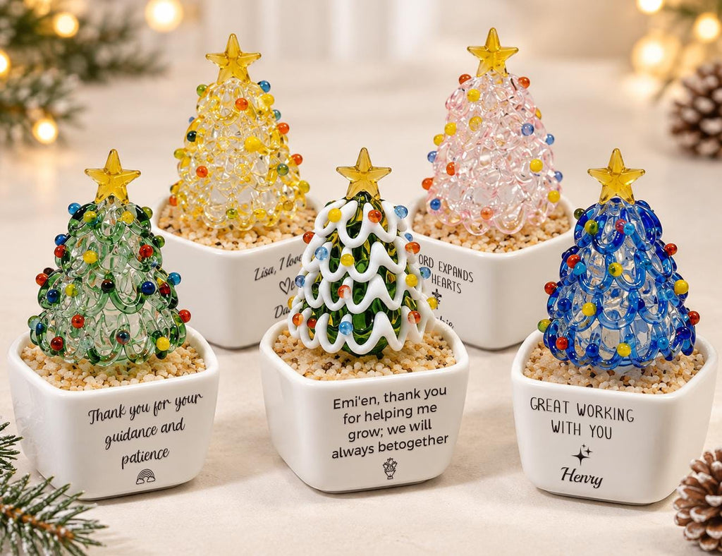May include: Five glass Christmas tree figurines in white square ceramic pots filled with sand. Each tree features a yellow star topper and colorful glass beads. The pots display various personalized messages and small graphic icons in black text.