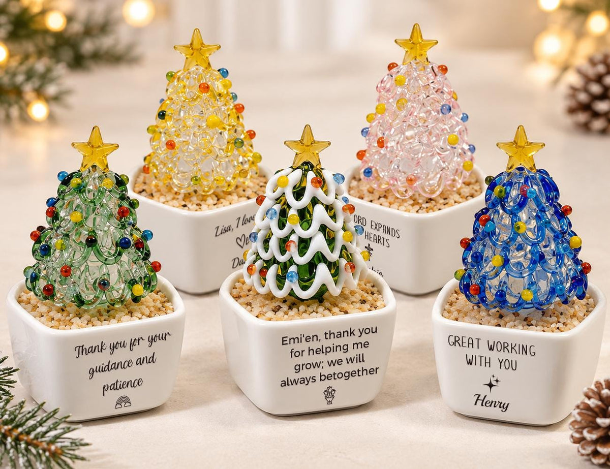 May include: Five glass Christmas tree figurines in white square ceramic pots filled with sand. Each tree features a yellow star topper and colorful glass beads. The pots display various personalized messages and small graphic icons in black text.