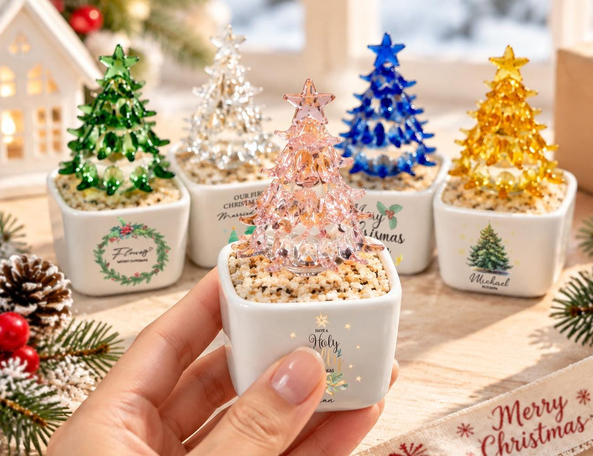 May include: A collection of five small, decorative glass Christmas trees in green, clear, pink, blue, and yellow, each topped with a star. The trees are set in square white ceramic pots filled with small pebbles, featuring various holiday-themed text.