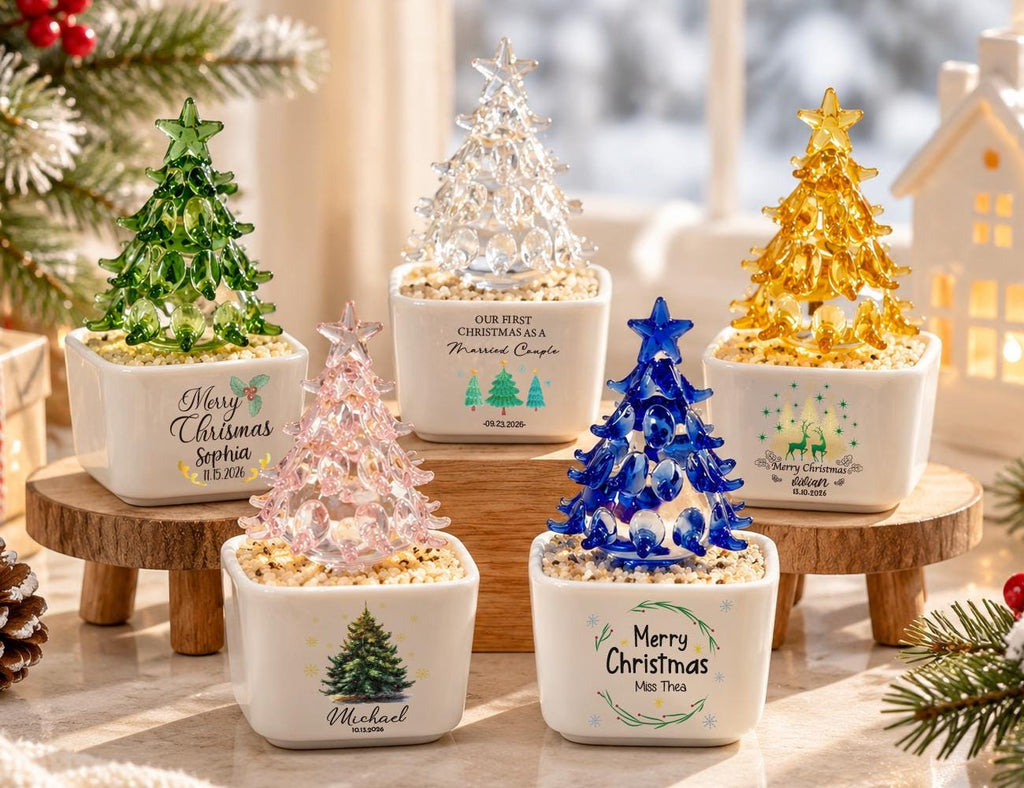 May include: Five glass Christmas tree figurines in white square ceramic pots filled with small stones. The trees are green, clear, yellow, pink, and blue. Each pot features custom text including names, dates, and phrases like Merry Christmas.