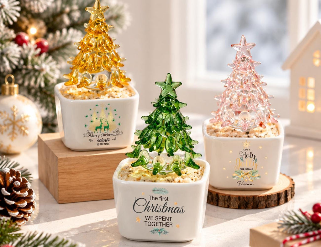May include: Three glass Christmas tree figurines in white square ceramic pots. One tree is yellow, one green, and one pink, each topped with a star. The pots feature printed text including "Merry Christmas," "The first Christmas WE SPENT TOGETHER," and "Have a Holy Christmas."