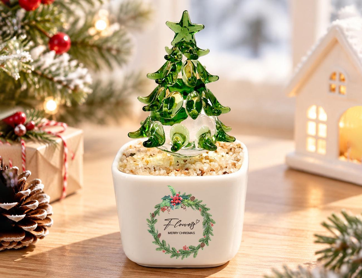 May include: A green glass Christmas tree figurine with a star topper, set in a white square ceramic pot filled with light-colored pebbles. The pot features a printed wreath graphic with the text "Flowers" and "MERRY CHRISMAS" in black ink.