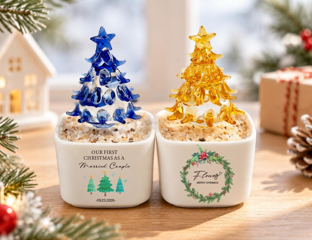 May include: Two square white ceramic planters filled with light-colored pebbles, each topped with a glass Christmas tree. The left planter features text reading "OUR FIRST CHRISTMAS AS A Married Couple" with three green trees and the date "09.23.2026". The right planter displays a wreath graphic with the text "Flower" and "MERRY CHRISTMAS".