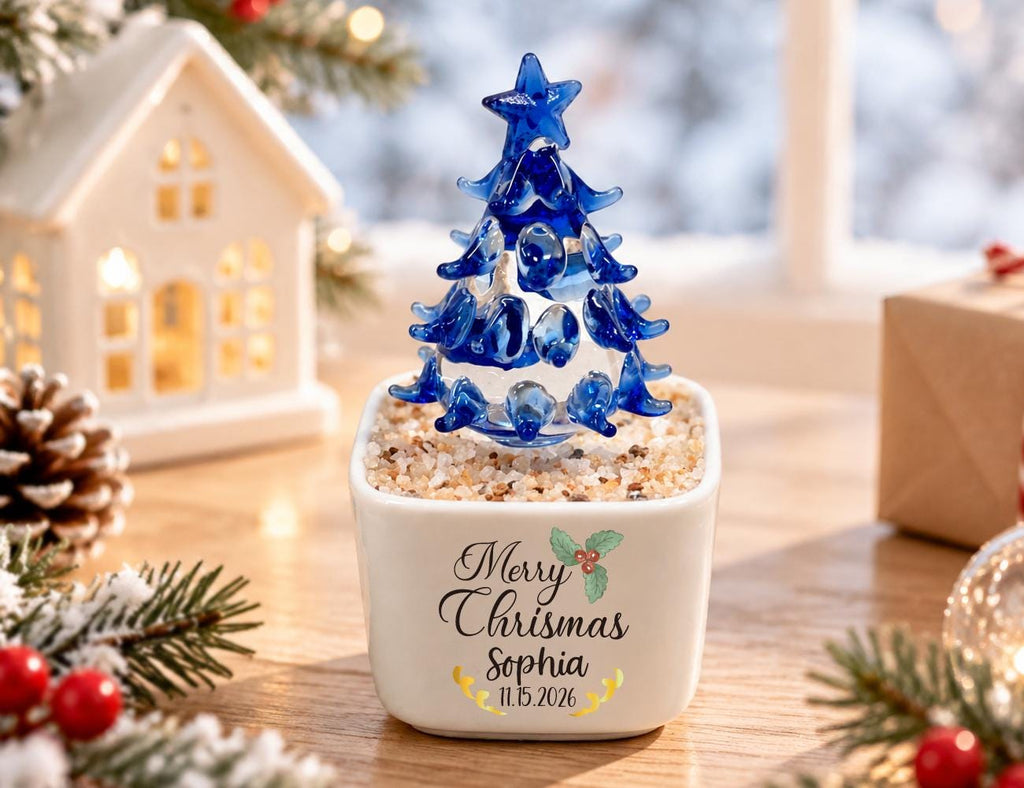 May include: A small blue glass Christmas tree sculpture with a star topper, set in a square white ceramic planter filled with light-colored pebbles. The planter features the text Merry Chrismas, the name Sophia, and the date 11.15.2026 in black.