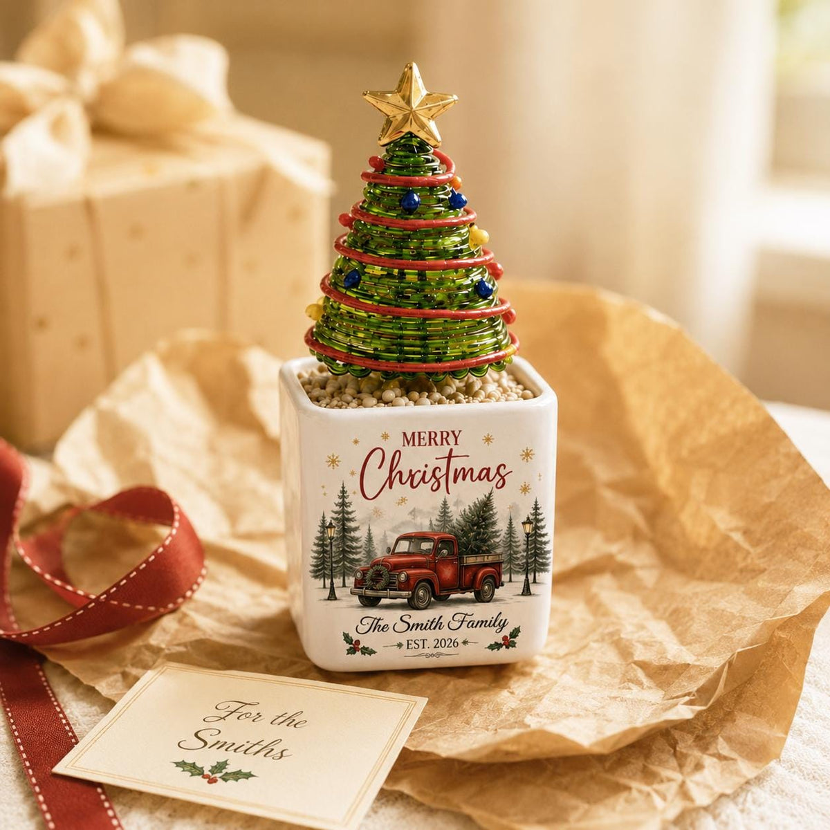May include: A decorative tabletop Christmas tree made of green wire and beads with a gold star topper, set in a white square ceramic planter. The planter features a red truck graphic, pine trees, and the text Merry Christmas The Smith Family EST. 2026.