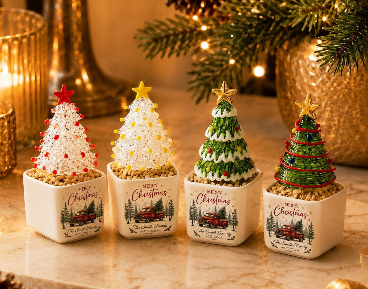May include: Four small decorative Christmas trees in square white ceramic pots. Each pot features a red truck graphic and the text Merry Christmas The Smith Family EST 2024. The trees have varied textures and colors, topped with stars.