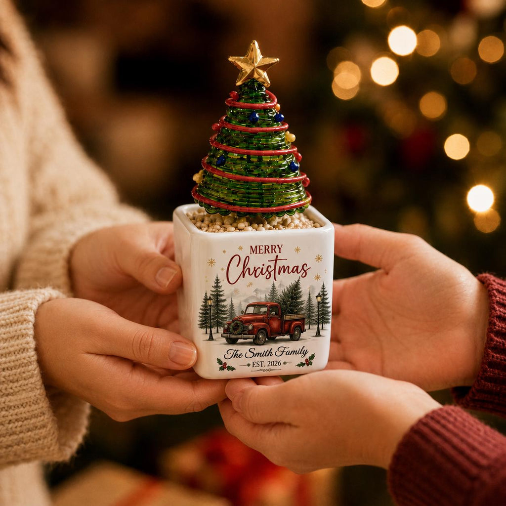 May include: A square white ceramic planter featuring a red truck graphic, pine trees, and the text Merry Christmas The Smith Family EST. 2026. A green wire Christmas tree with small colorful ornaments and a gold star topper sits inside the planter.