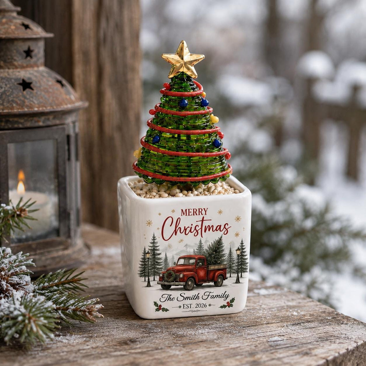 May include: A white square ceramic planter featuring a red truck graphic, the text Merry Christmas, and The Smith Family EST. 2026. A green wire-wrapped Christmas tree with small colorful beads and a gold star topper sits inside the planter.