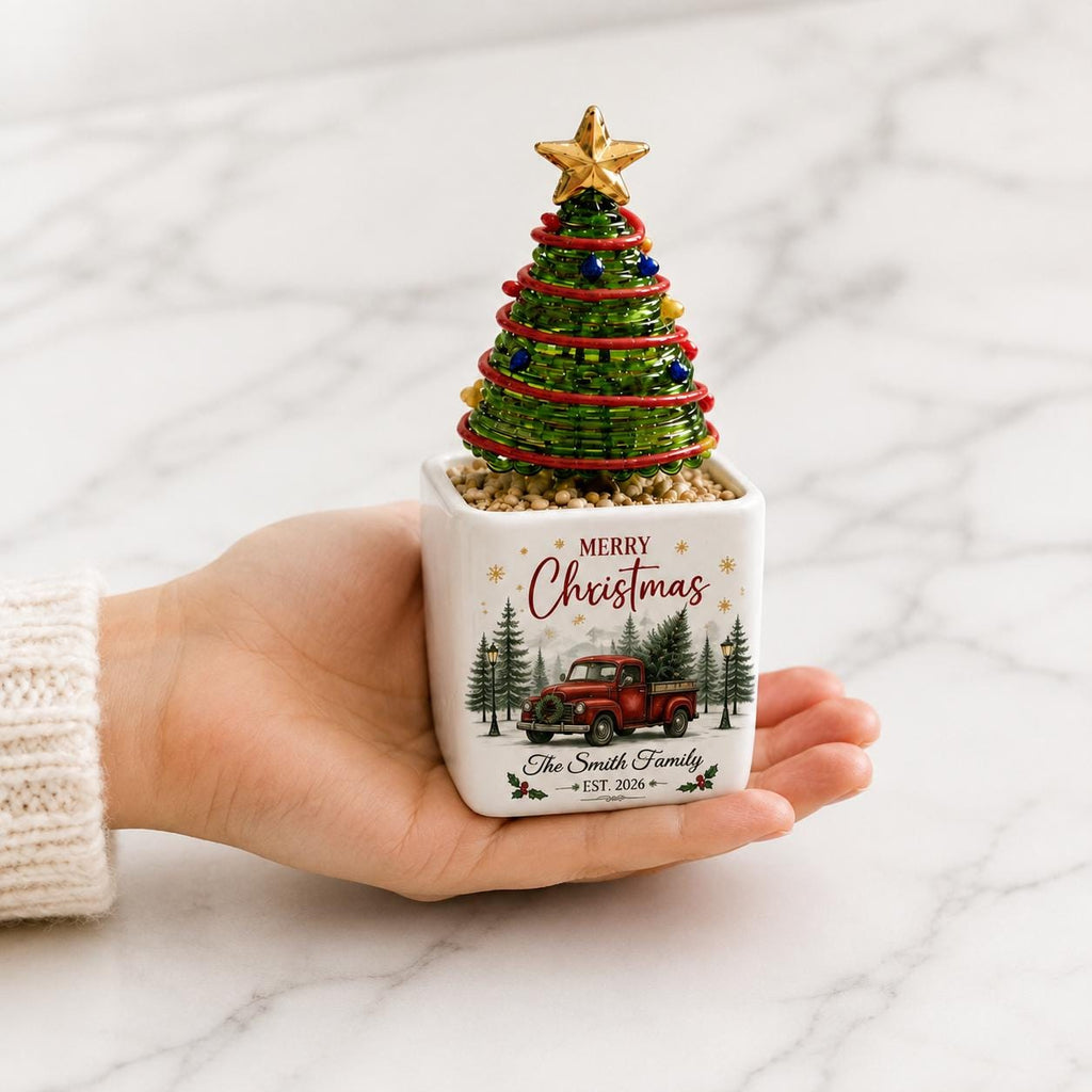 May include: A square white ceramic planter featuring a red truck graphic, pine trees, and the text Merry Christmas The Smith Family EST. 2026. A small green wire Christmas tree with colorful beads and a gold star topper sits inside the planter.