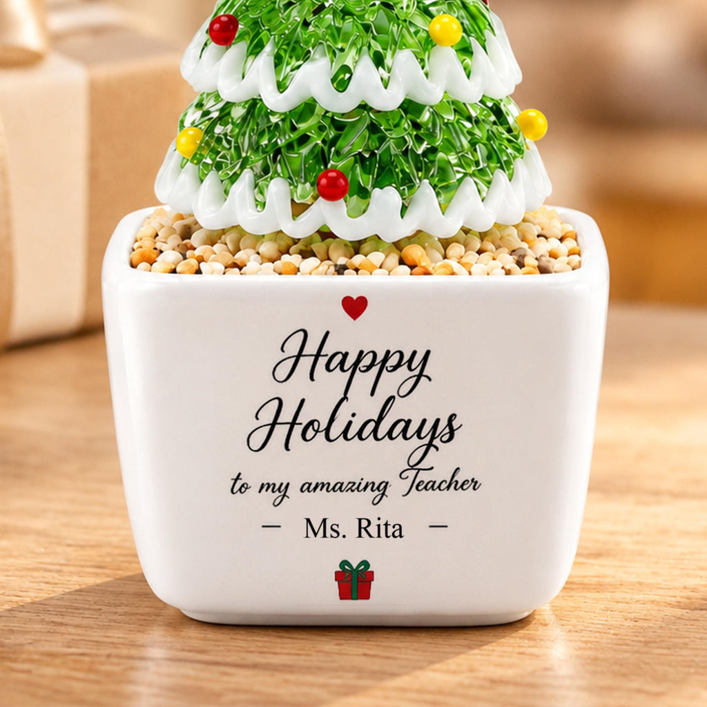 May include: A white square ceramic planter filled with small pebbles and a green glass Christmas tree ornament with white trim and colorful beads. The planter features the text Happy Holidays to my amazing Teacher - Ms. Rita - and a small red gift icon.