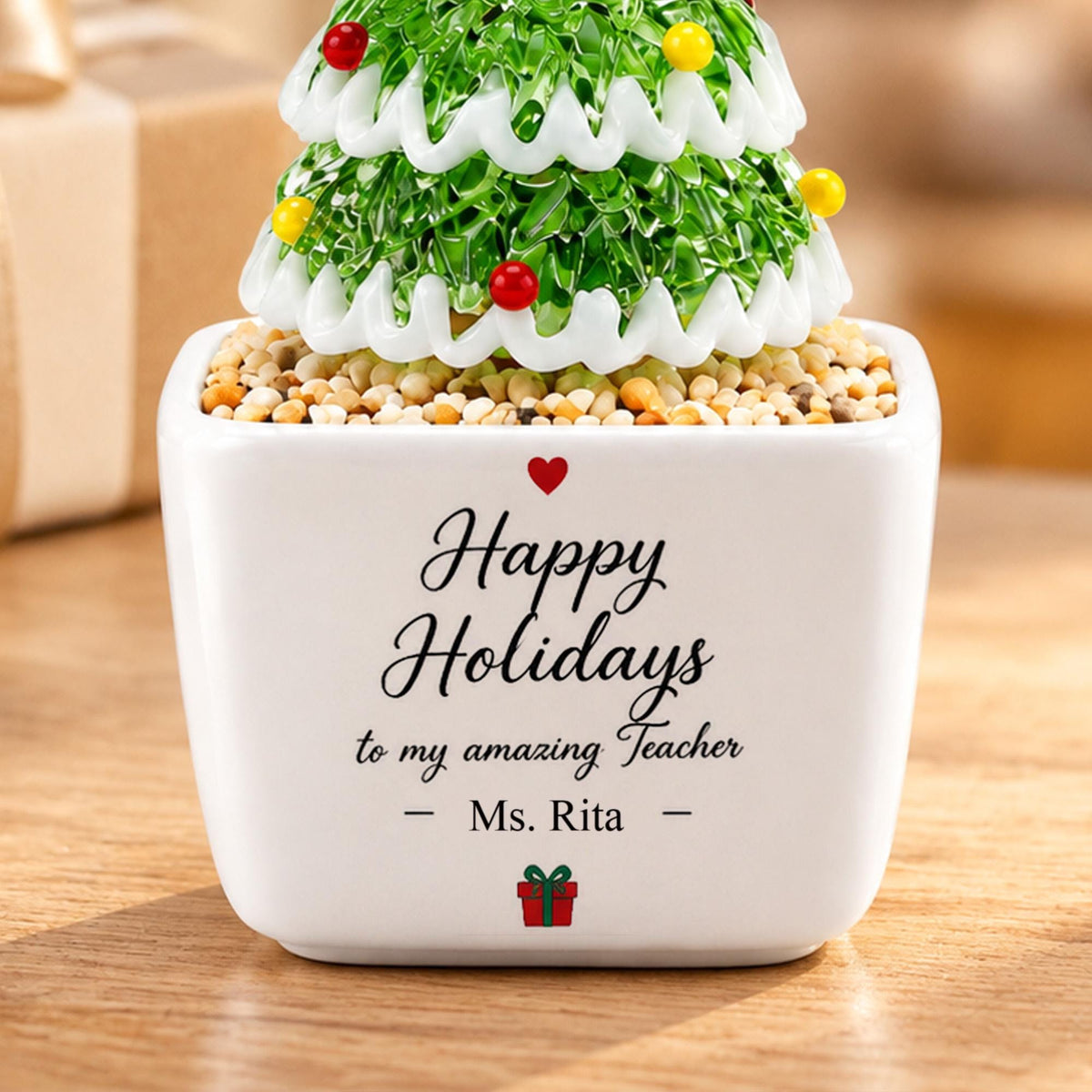 May include: A white square ceramic planter filled with small pebbles and a green glass Christmas tree ornament with white trim and colorful beads. The planter features the text Happy Holidays to my amazing Teacher - Ms. Rita - and a small red gift icon.