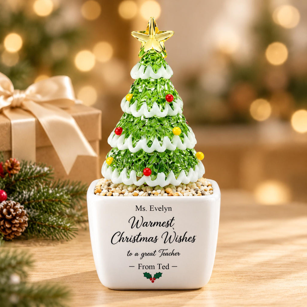 May include: A decorative glass Christmas tree ornament with green foliage, white snow-like trim, and small red and yellow spheres, topped with a gold star. It sits in a white square planter with the text Ms. Evelyn Warmest Christmas Wishes to a great Teacher - From Ted - and a small holly icon.