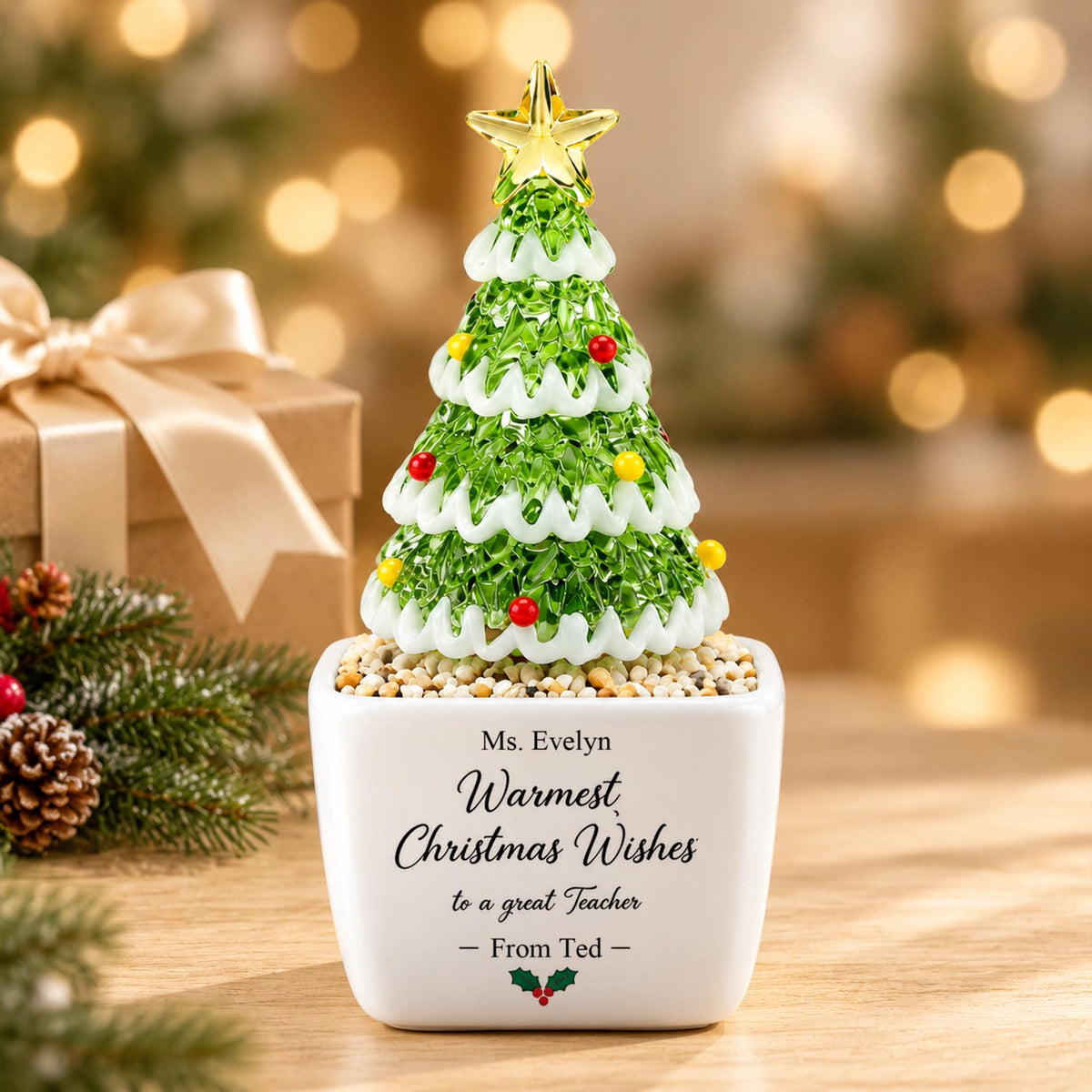May include: A decorative glass Christmas tree ornament with green foliage, white snow-like trim, and small red and yellow spheres, topped with a gold star. It sits in a white square planter with the text Ms. Evelyn Warmest Christmas Wishes to a great Teacher - From Ted - and a small holly icon.