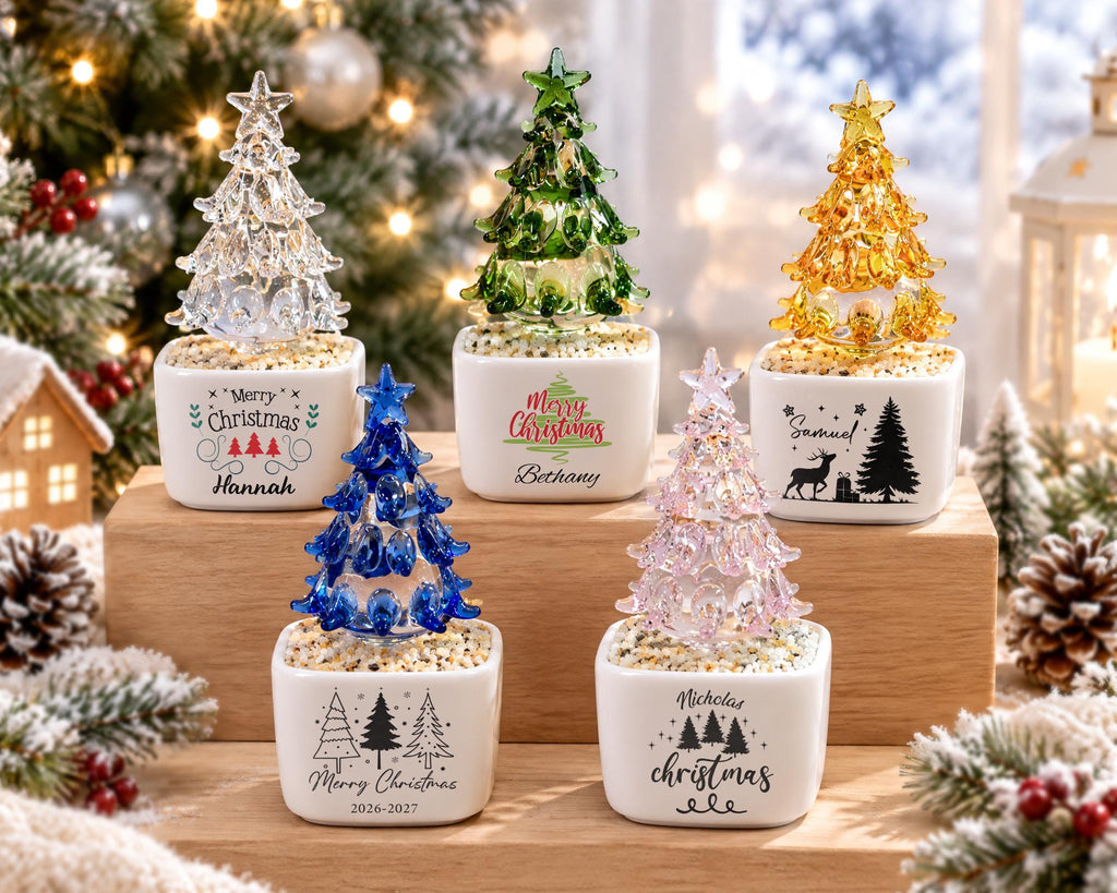 May include: Five glass Christmas tree figurines in white ceramic pots filled with small stones. The trees are clear, green, yellow, blue, and pink. Each pot features unique text and graphics, including names like Hannah, Bethany, Samuel, and Nicholas.