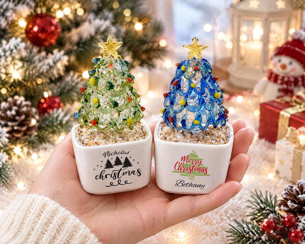 May include: Two small glass Christmas tree ornaments in white square ceramic pots. One tree is green with yellow and red accents, labeled Nicholas Christmas. The other is blue with yellow and red accents, labeled Merry Christmas Bethany.
