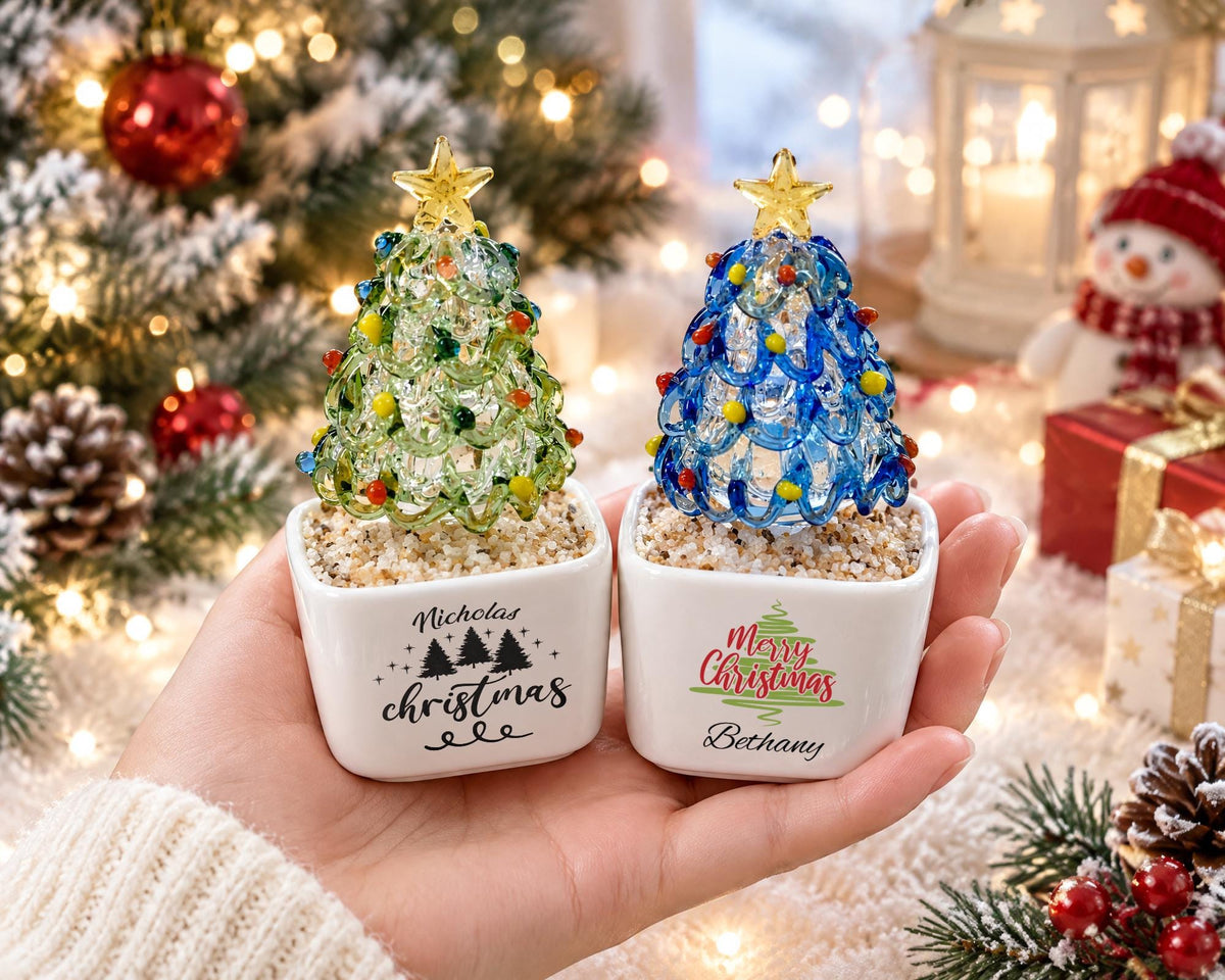 May include: Two small glass Christmas tree ornaments in white square ceramic pots. One tree is green with yellow and red accents, labeled Nicholas Christmas. The other is blue with yellow and red accents, labeled Merry Christmas Bethany.