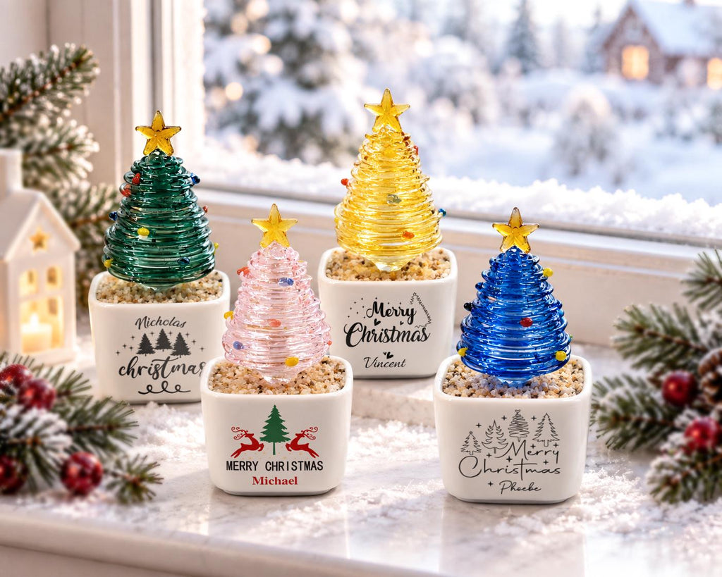 May include: Four glass Christmas tree ornaments in white ceramic pots filled with pebbles. The trees are green, pink, yellow, and blue, each topped with a yellow star. The pots feature printed text including names like Nicholas, Vincent, Michael, and Phoebe.