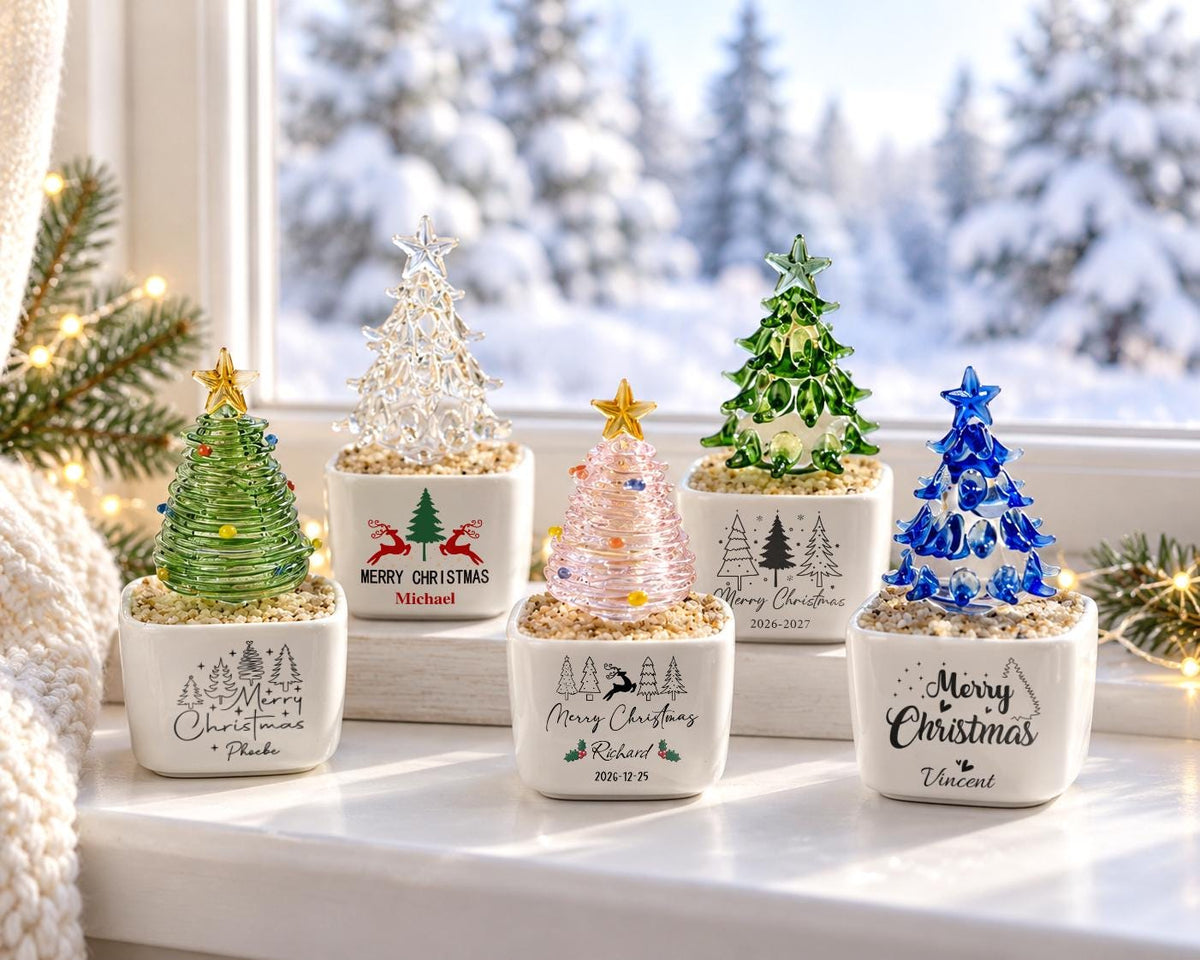May include: Five small glass Christmas trees in white square ceramic pots filled with decorative sand. Each tree features a different color: green, clear, pink, green, and blue, topped with a star. The pots display Merry Christmas text.