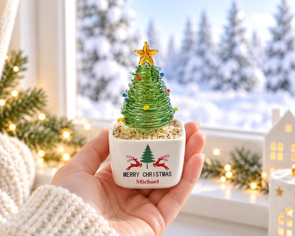 May include: A small glass Christmas tree figurine with a yellow star topper and colorful glass beads, set in a white square ceramic planter filled with light-colored pebbles. The planter features a green tree, red reindeer, and the text Merry Christmas Michael.