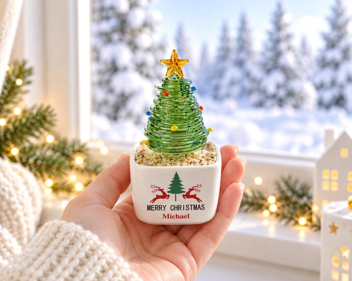 May include: A small glass Christmas tree figurine with a yellow star topper and colorful glass beads, set in a white square ceramic planter filled with light-colored pebbles. The planter features a green tree, red reindeer, and the text Merry Christmas Michael.