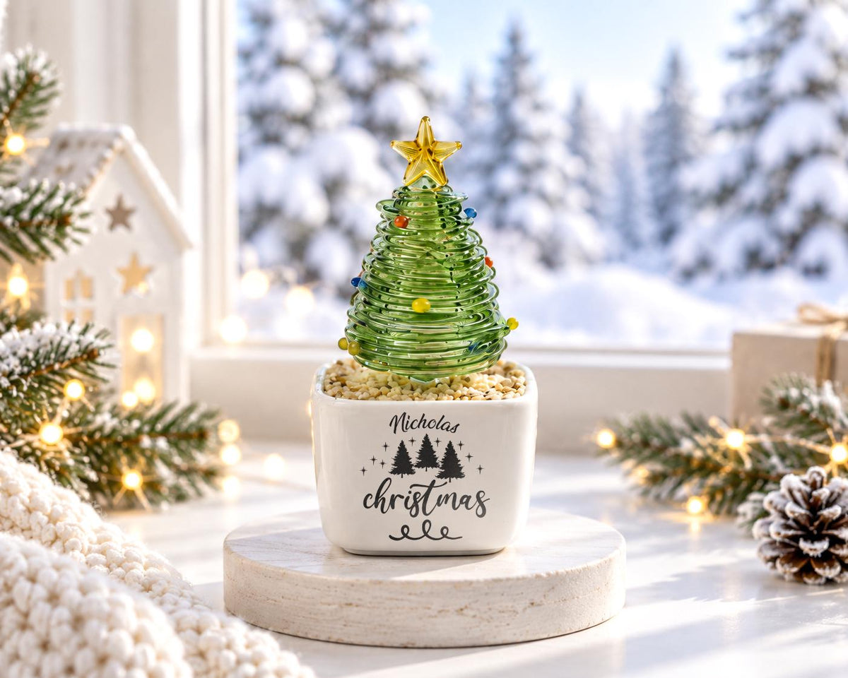 May include: A green glass spiral Christmas tree ornament with a yellow star topper, set in a white square ceramic planter filled with small pebbles. The planter features the black text Nicholas christmas and a graphic of three pine trees.