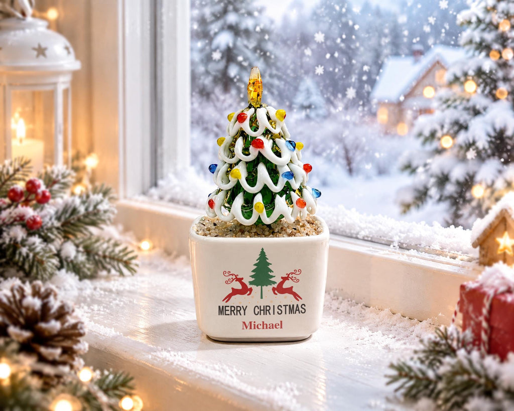 May include: A glass Christmas tree figurine with white swirled branches and colorful glass bulbs, topped with a yellow star. The tree sits in a square white ceramic pot featuring a green tree, two red reindeer, and the text Merry Christmas Michael.