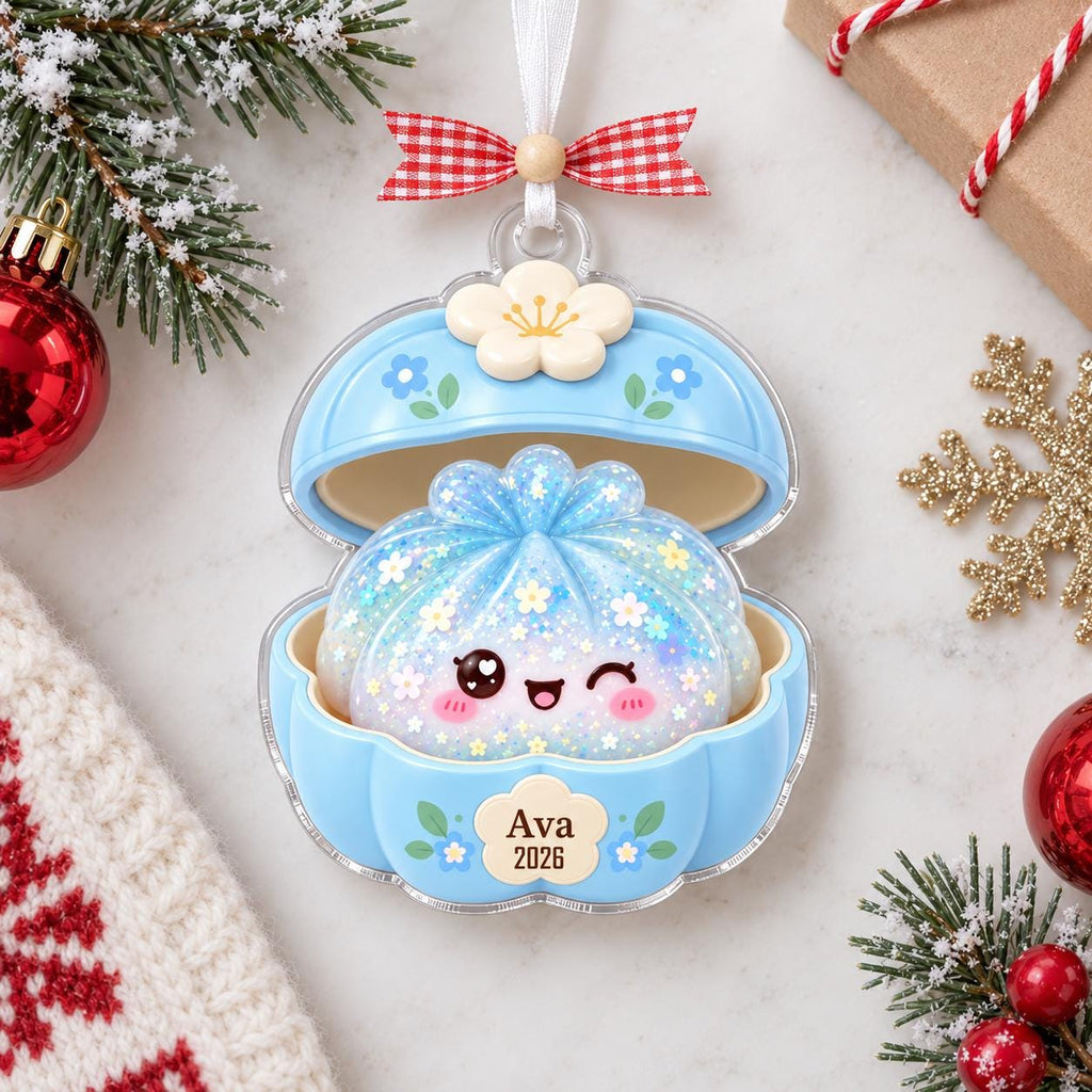 May include: A light blue clam-shaped ornament featuring a glittery, smiling dumpling character inside. The exterior is decorated with small blue flowers and a white flower topper. A cream-colored plaque on the front displays the text Ava 2026.