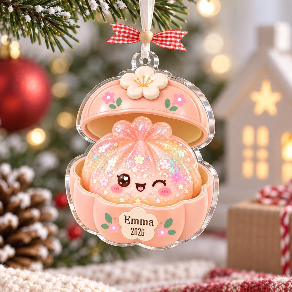 May include: A peach-colored, clam-shaped Christmas ornament featuring a cute, winking dumpling character with iridescent glitter and floral accents. The shell is topped with a white flower and includes the printed text Emma 2026 on the front.