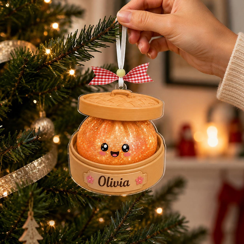 May include: A Christmas tree ornament shaped like a dumpling in a bamboo steamer basket. The orange, glittery dumpling has a smiling face. The base features the name Olivia with small pink flowers. A red and white gingham bow hangs above.