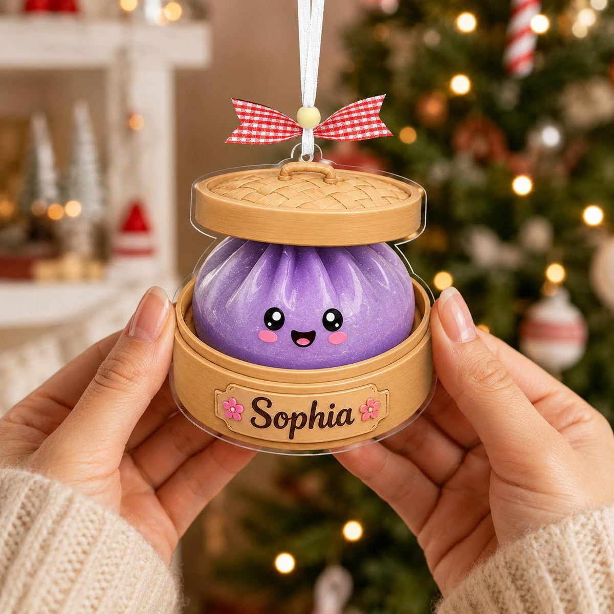 May include: A hanging ornament shaped like a purple dumpling with a smiling face inside a bamboo steamer basket. The base features a label with the name Sophia and small pink flower icons. A red and white checkered ribbon is tied at the top.