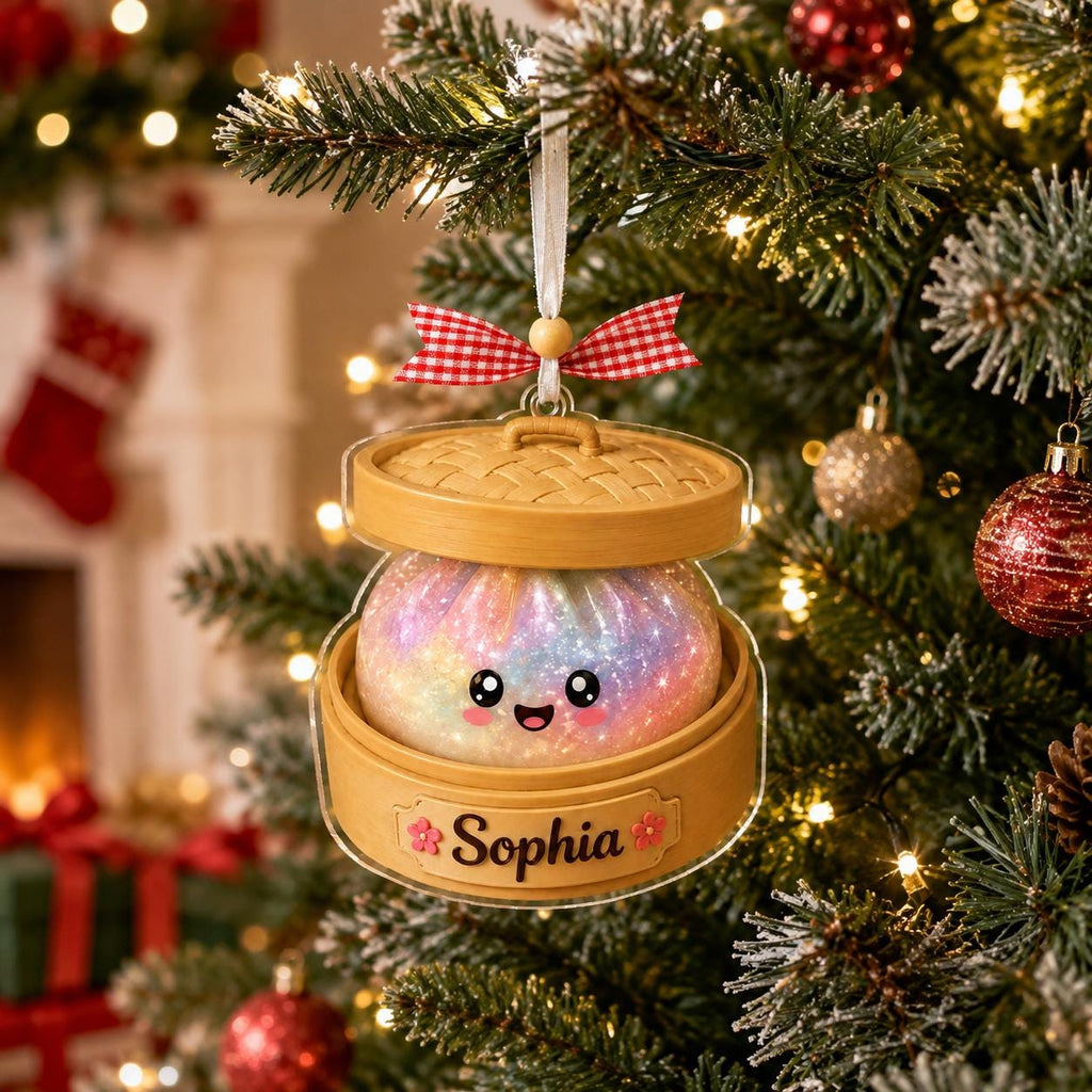 May include: A Christmas tree ornament shaped like a bamboo steamer basket containing a round, iridescent, glittery dumpling with a smiling face. The base features a tan label with the name Sophia and two small pink flowers. A red and white gingham bow sits on top.