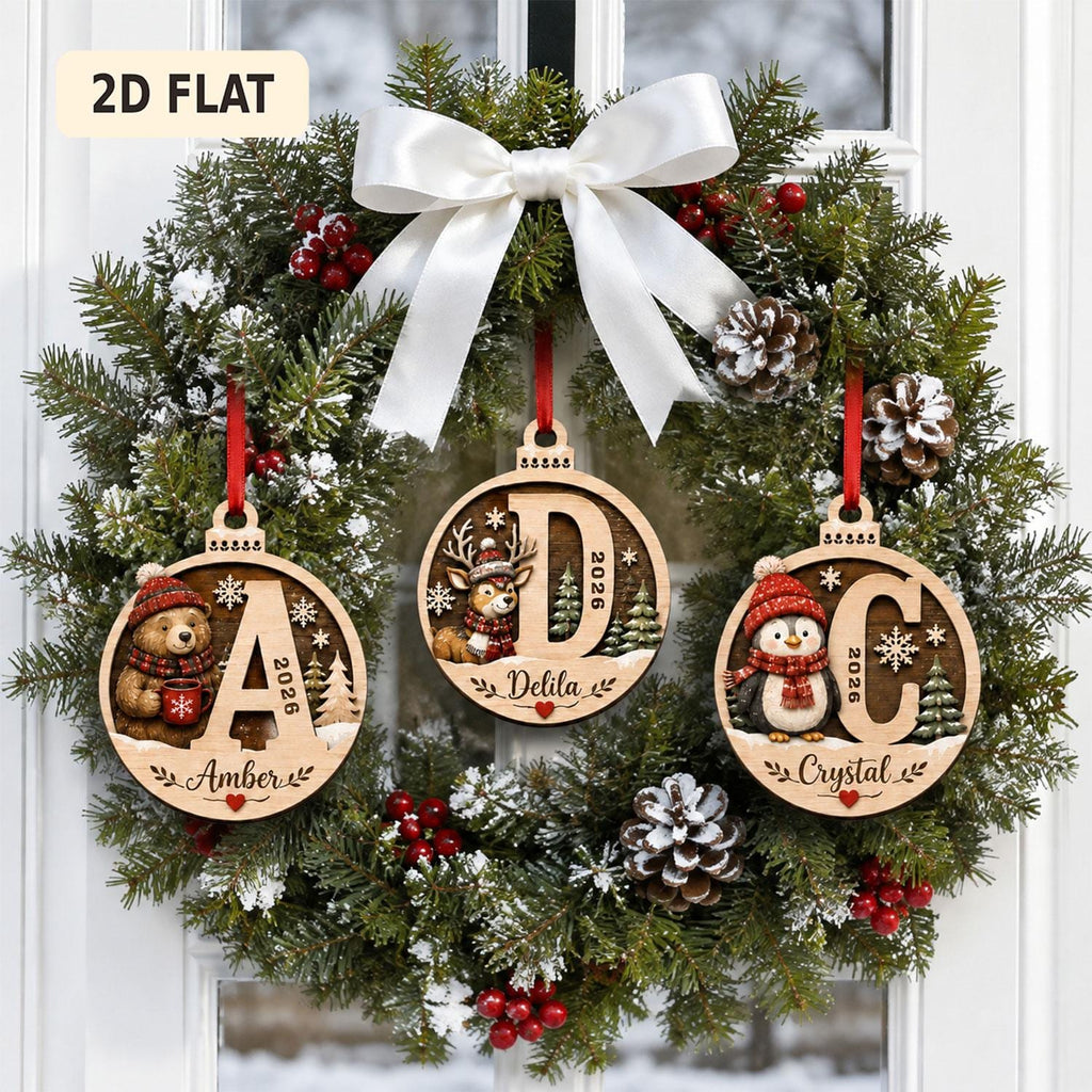 May include: Three circular wooden Christmas ornaments hanging on a green wreath with a white bow. Each ornament features a large letter, the year 2026, a name, and a festive character: a bear with the letter A, a deer with D, and a penguin with C.